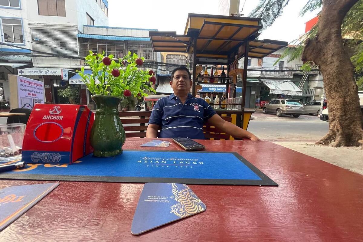 A man seated at a table with a blue placemat, a red and blue object, and a green vase with red flowers.