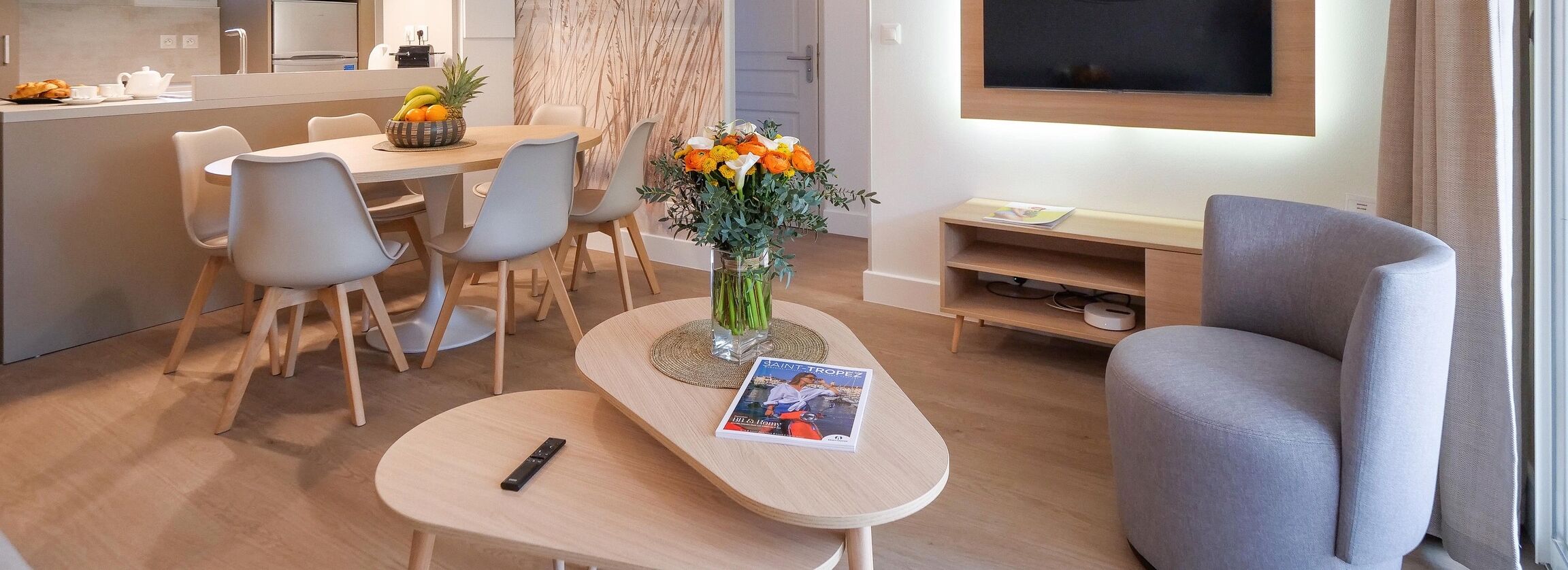 A modern living room with a dining area, a TV mounted on the wall, and a remote control on a small table.