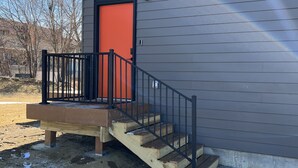 A building with a grey exterior, a red door, and a wooden staircase leading to a porch with a black railing. - OranGe DeLuxe - No Cleaning Fee, Centrally Located. (Omaha)
