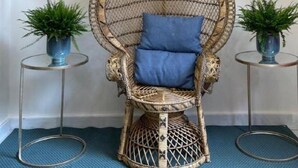 Una silla de mimbre con un cojín azul en una habitación con suelo azul, un espejo grande y dos plantas en macetas.