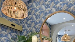 Una habitación con papel tapiz estampado, un espejo redondo, un sofá con cojines y dos mesas de centro de mimbre con plantas.