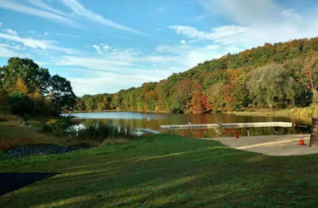 Eine friedliche Seenlandschaft mit einem hölzernen Steg, der ins Wasser führt, umgeben von üppiger Vegetation und Bäumen im Herbstlaub.