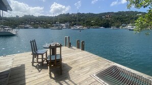 Un muelle de madera con una mesa y sillas, con vistas a una marina con varios barcos y un paisaje montañoso al fondo.
