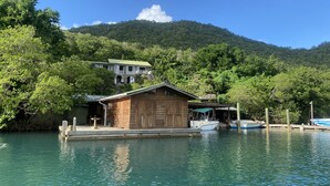 Un muelle de madera con una cabaña pequeña, barcos y una casa en una colina bajo un cielo azul despejado.