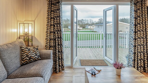 A cozy living room with a sofa, a coffee table, and a view of a green lawn and houses through large glass doors. - 2 bedroom accommodation in Willington, near Derby (Willington, near Derby)