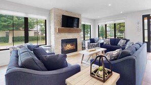 A modern living room with a fireplace, large windows, and a sectional sofa. - ExpERIEnce South Haven | Beach Farmhouse Steps to South Beach and Lake Michigan (South Haven)
