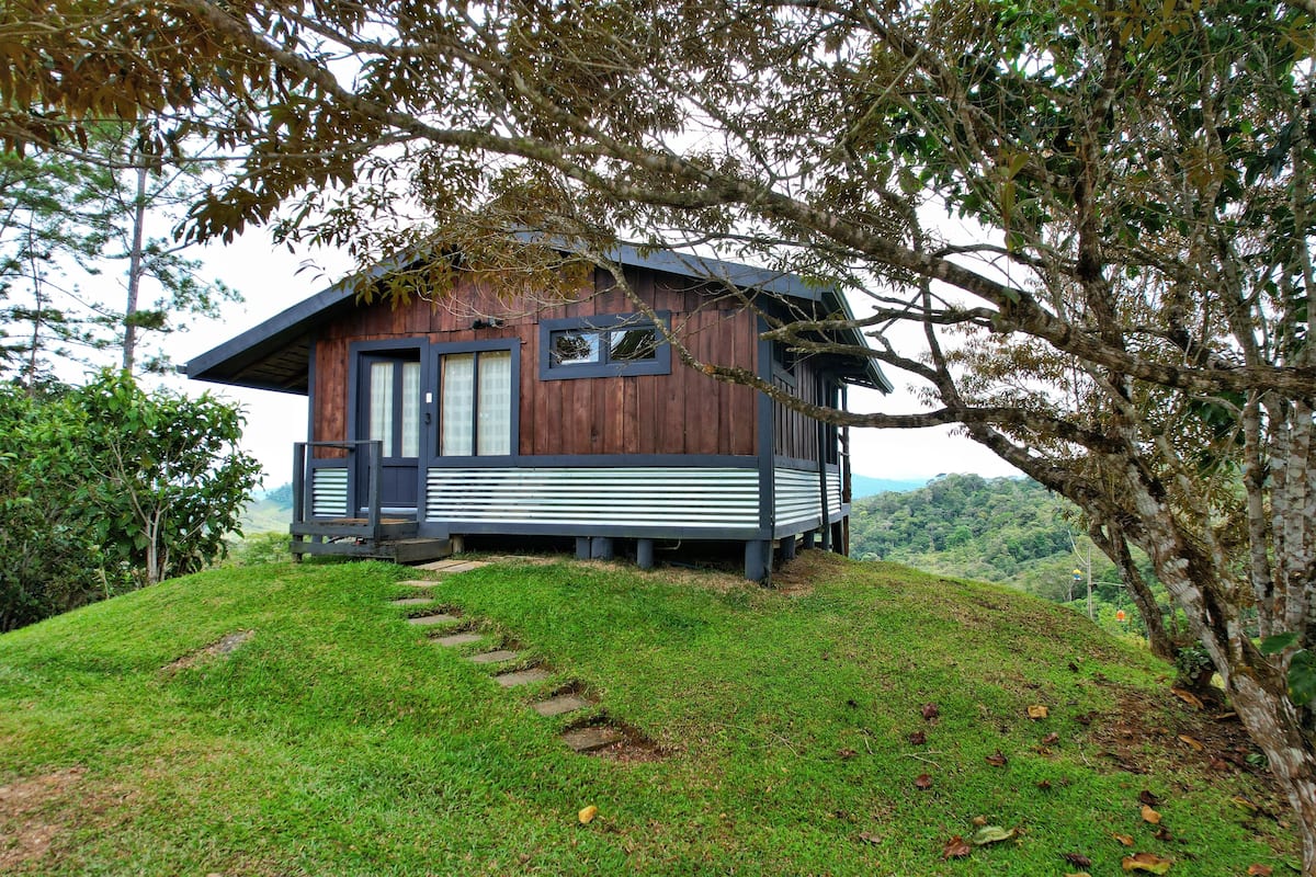 A wooden cabin with a porch, surrounded by lush greenery and hills.
