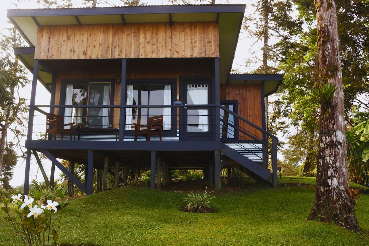 A two-story wooden cabin with a balcony, surrounded by greenery.