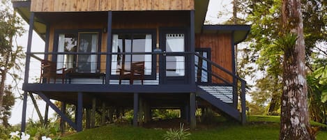 A two-story wooden cabin with a balcony, surrounded by greenery.