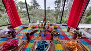 A dining table set with a colorful tablecloth, various dishes, and drinks, with a scenic view through large windows.