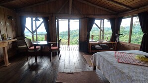 A wooden cabin with a dining area, a bed, and large windows offering a view of the forest.
