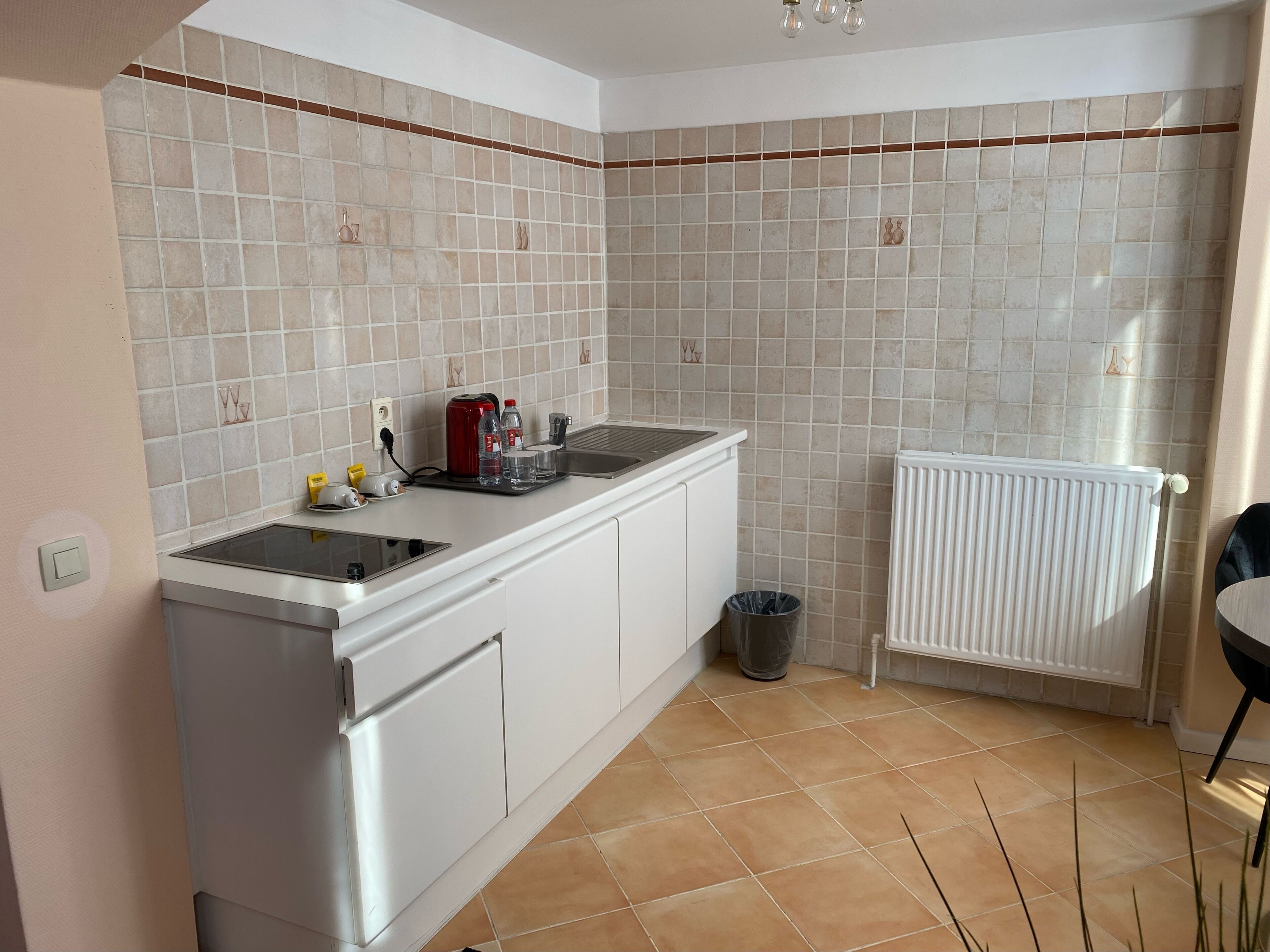 A kitchen with a white countertop, a red kettle, a trash can, and a white radiator.
