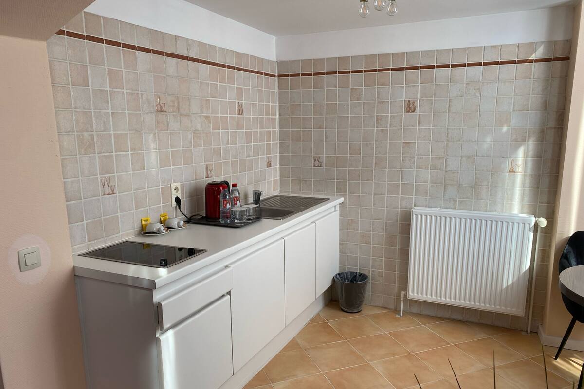 A kitchen with a white countertop, a red kettle, a trash can, and a white radiator.