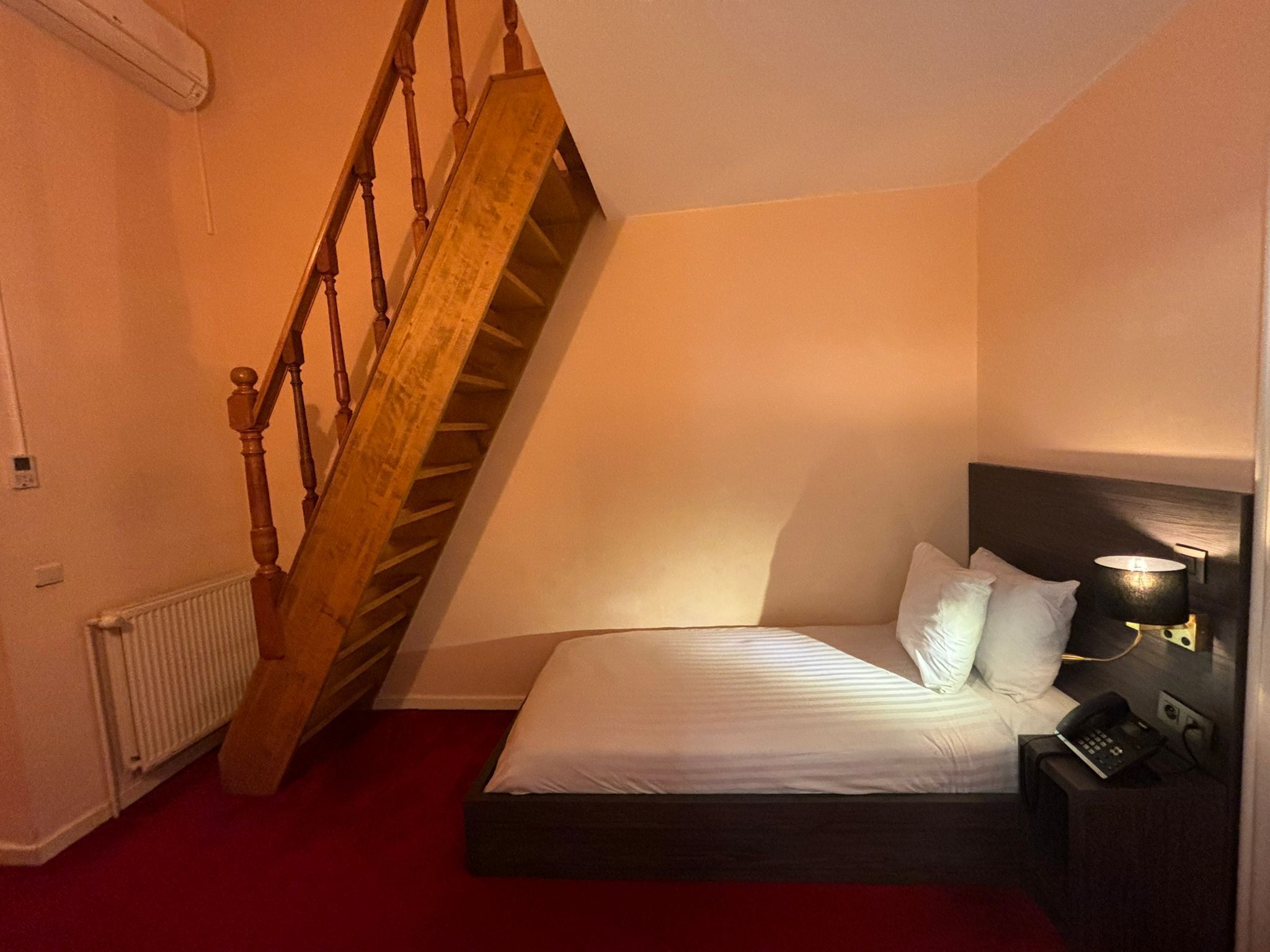 A bedroom with a bed, a nightstand, a lamp, a telephone, a radiator, and a wooden staircase.