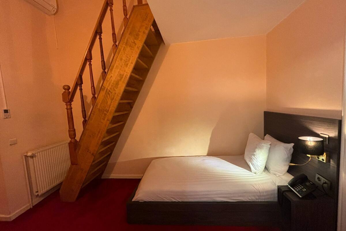A bedroom with a bed, a nightstand, a lamp, a telephone, a radiator, and a wooden staircase.