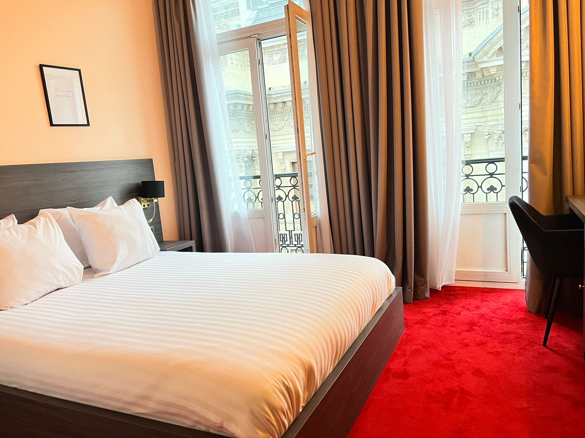 A hotel room with a bed, a desk, a chair, and a balcony with curtains.