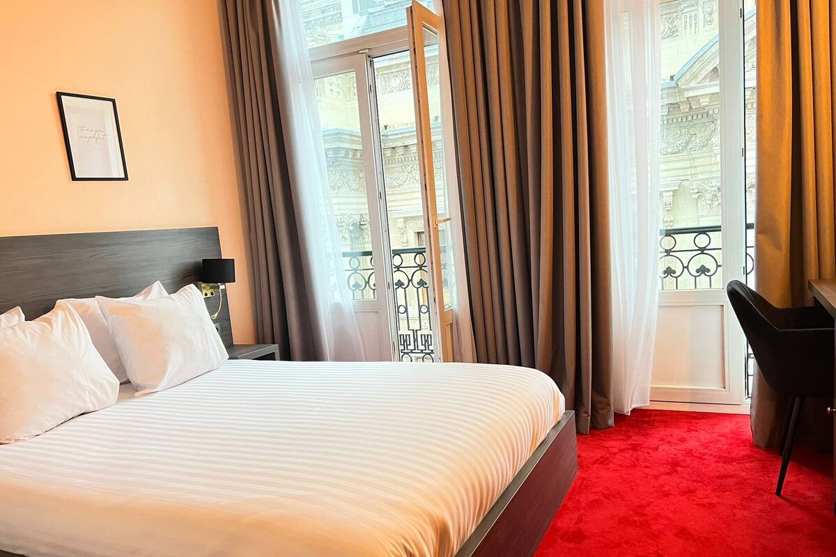 A hotel room with a bed, a desk, a chair, and a balcony with curtains.