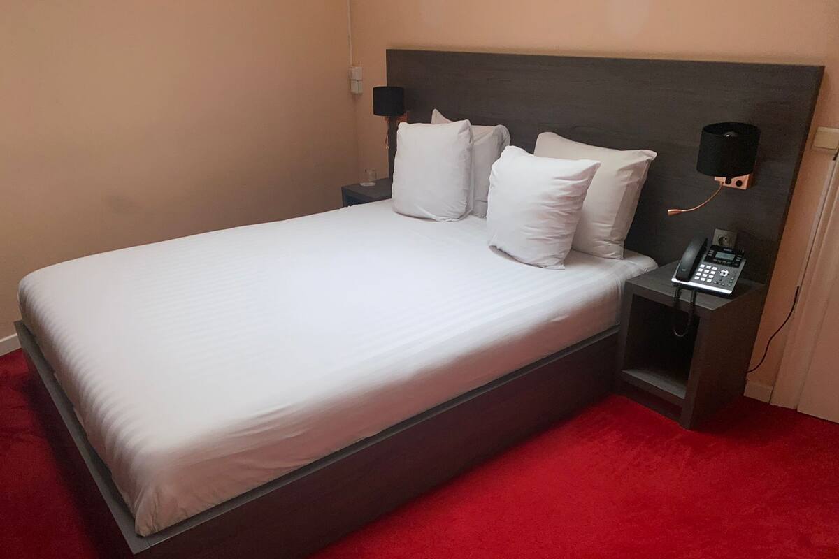 A single bed with white linens, a bedside telephone, and a nightstand with a lamp.