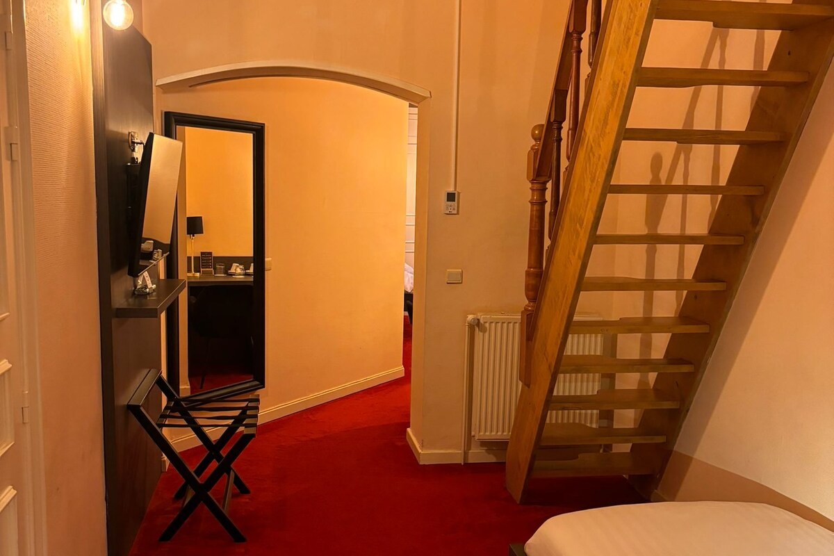 A hotel room with a red carpet, a bed, a wooden ladder, a desk with a mirror, and a window with curtains.