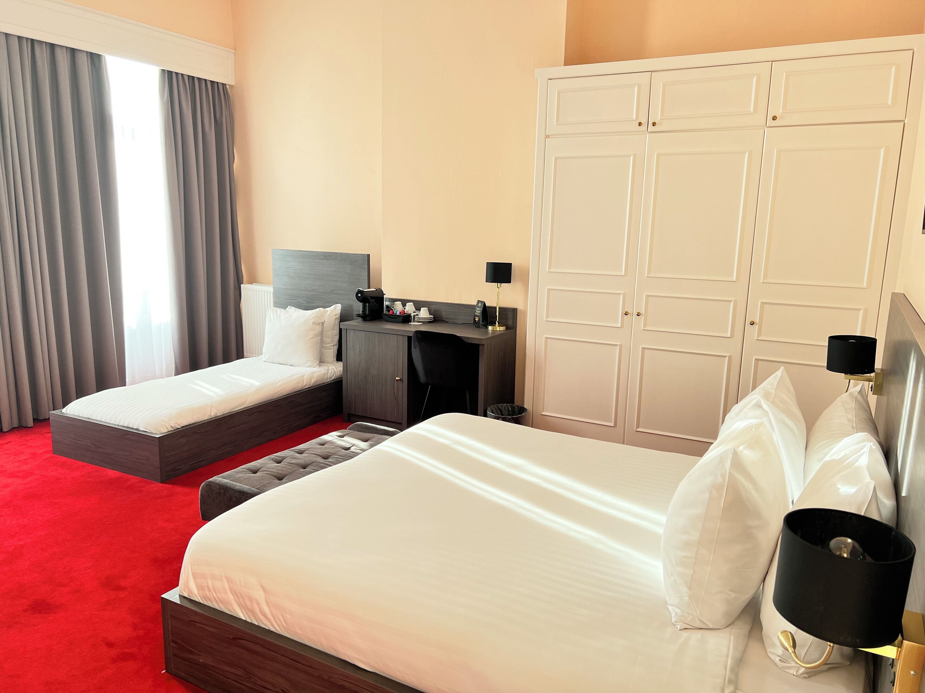 A hotel room with two beds, a desk, a wardrobe, and a window with curtains.