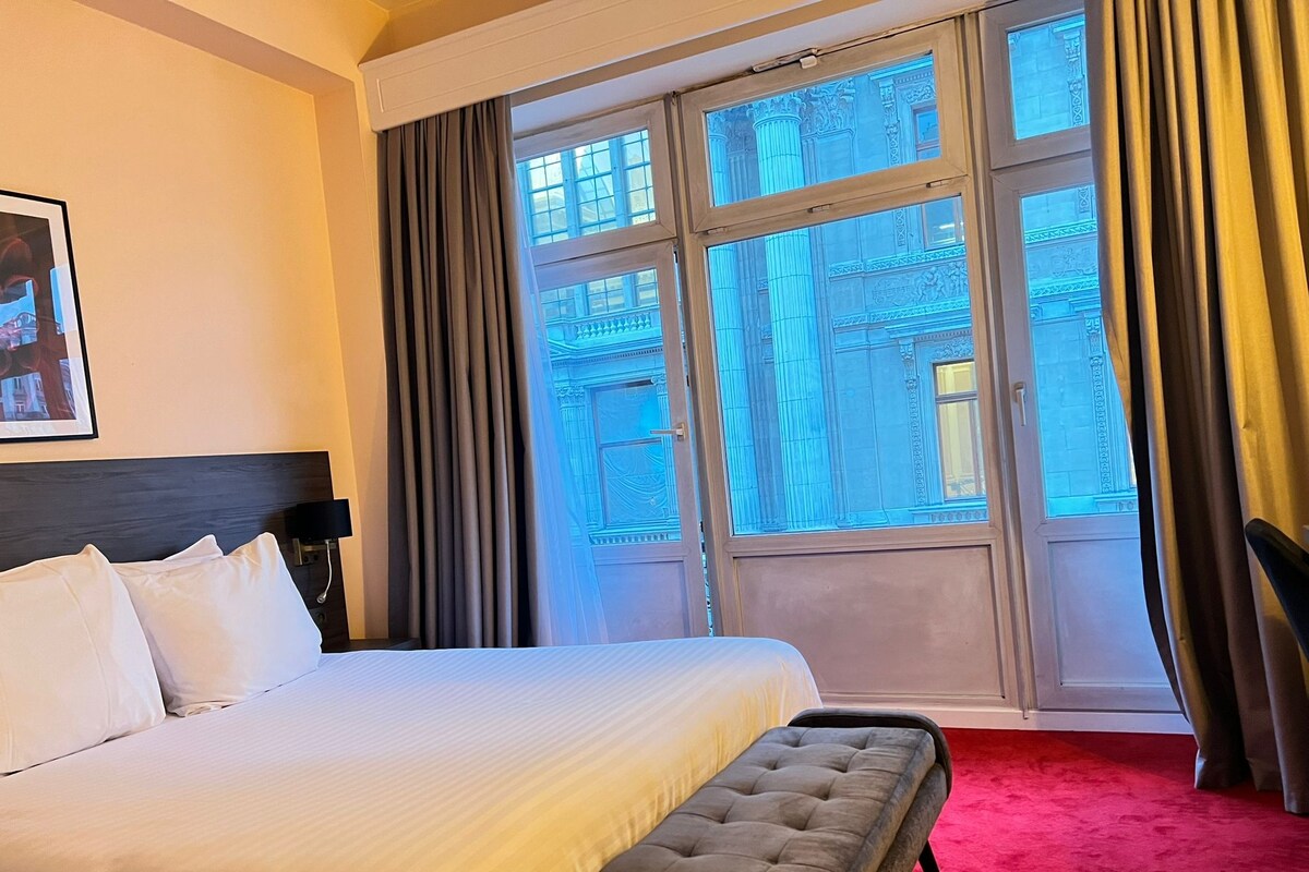 A hotel room with a large bed, a bench, a nightstand, and a view of a building through the window.