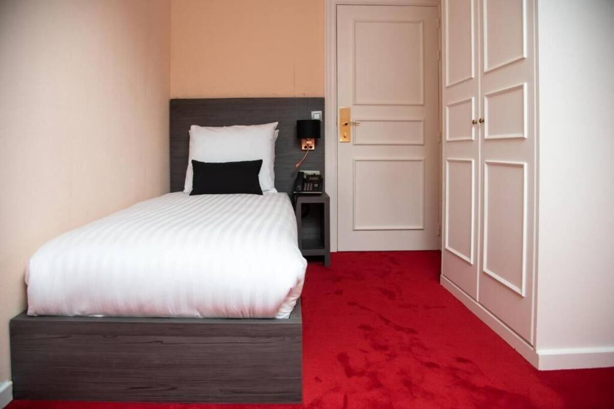 A neatly made bed with a wooden headboard, a bedside table with a phone, and a red carpeted floor.