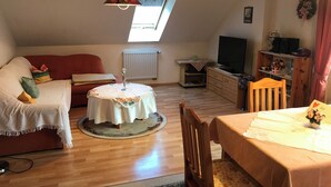 Een knus loft-achtig vertrek met een houten vloer, een ronde tafel met een witte tafelkleed, een rode bank en een televisie op een houten dressoir.