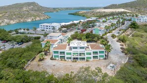 A coastal town with a prominent white building with green windows, surrounded by palm trees and a clear blue sea. - Electricity Included in rental price! (Willemstad)