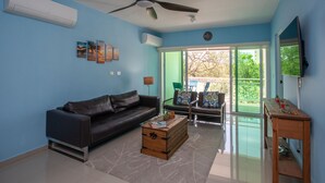 A living room with a dark brown sofa, a wooden coffee table, a ceiling fan, and a large window with a view of greenery. - Electricity Included in rental price! (Willemstad)