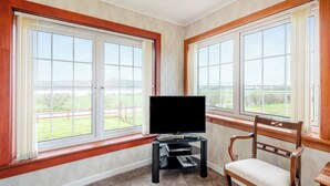 A room with large windows, a flat-screen TV on a stand, a wooden chair, and a carpeted floor. - 4 bedroom accommodation in Kingarth (Kingarth)