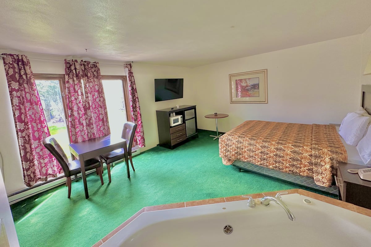 A hotel room with a bed, a desk, a chair, a TV, a small table, and a bathroom with a bathtub.