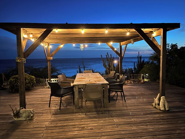 Une terrasse en bois avec une table et des chaises, située sous une pergola et offrant une vue sur l’océan.