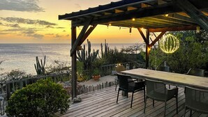 Een houten terras met een tafel en stoelen, uitkijkend op een wateroppervlak bij zonsondergang.