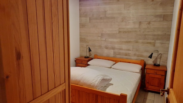 Una cama con estructura de madera, ropa de cama blanca, dos mesitas de noche con lámparas y una pared de madera.