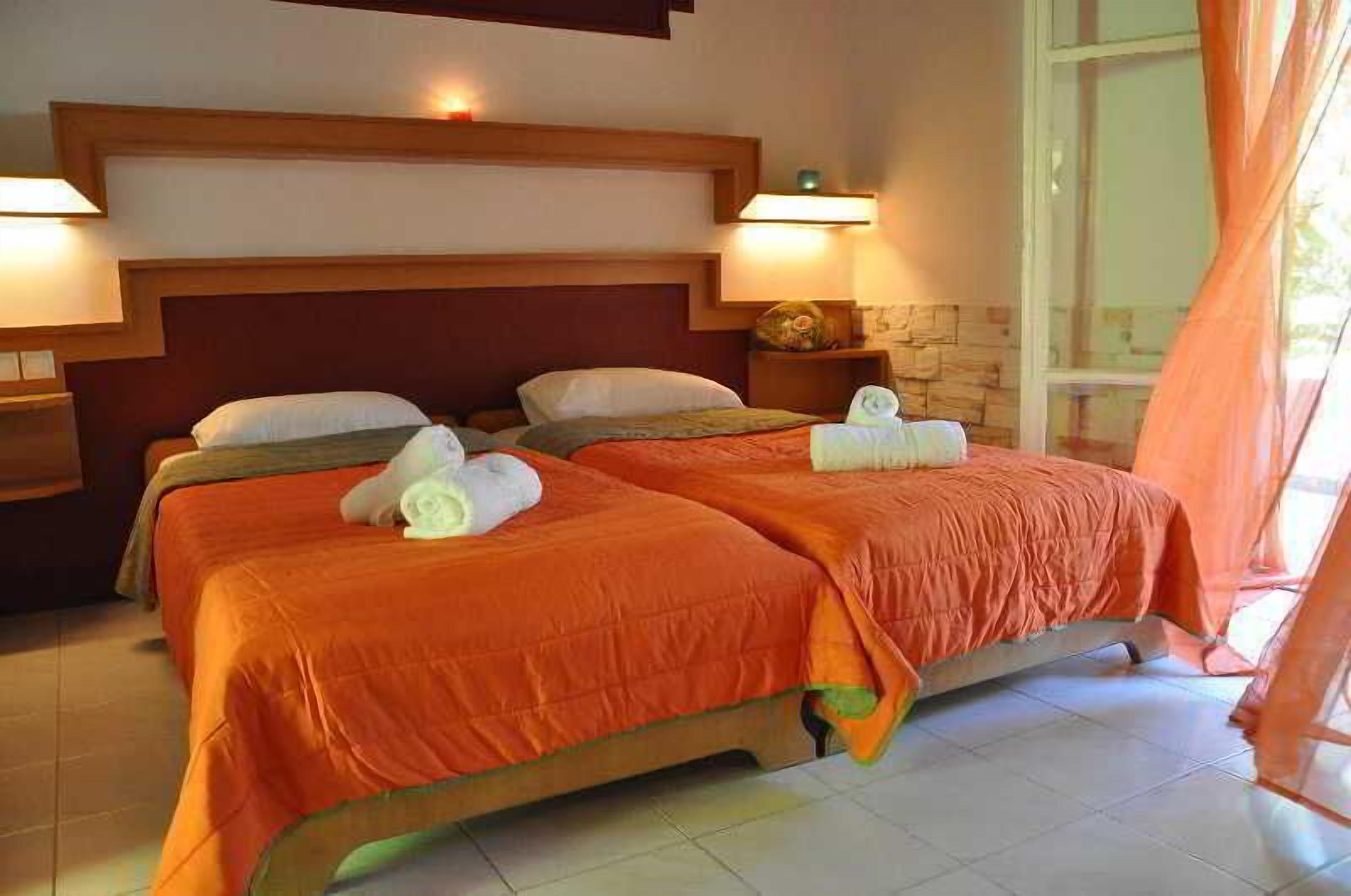 A hotel room with two beds, each with an orange bedspread and white pillows, a wooden headboard, and a nightstand with a lamp.
