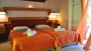 A hotel room with two beds, each with an orange bedspread and white pillows, a wooden headboard, and a nightstand with a lamp. - Tassos Apartments (Corfu)