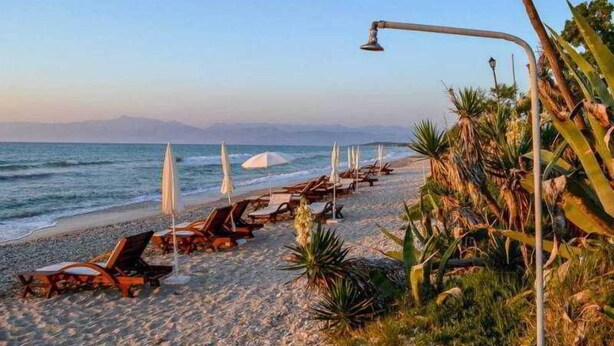A beach with lounge chairs and umbrellas, a clear sky, and a view of the sea and distant mountains. - Tassos Apartments (Corfu)