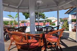A covered outdoor seating area with wicker chairs and a glass table, overlooking a pool and buildings. - Tassos Apartments (Corfu)