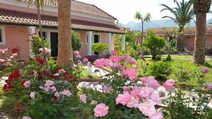 A building with a tiled roof and a garden with pink and red flowers. - Tassos Apartments (Corfu)