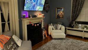 En stue med pejs, et tv ophængt over, og et hyggeligt opholdsområde med sofa og skammel.