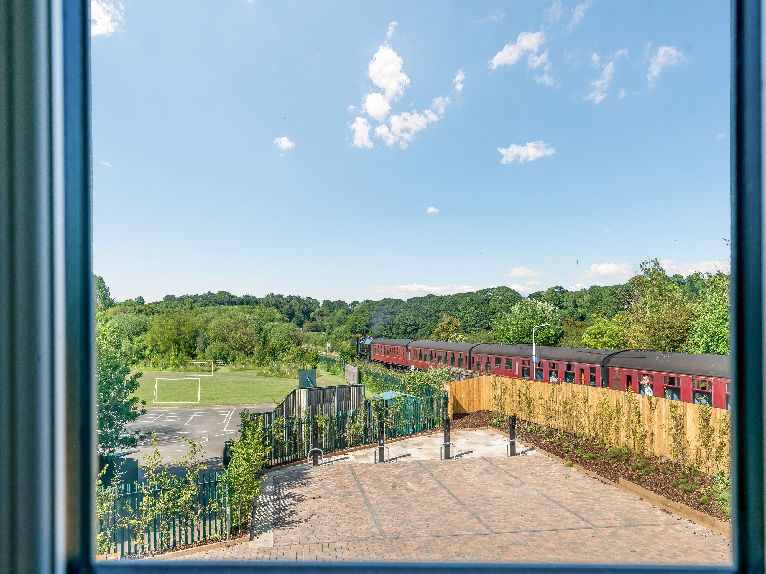 A train station with a red train, a sports field, and a forested hill in the background.