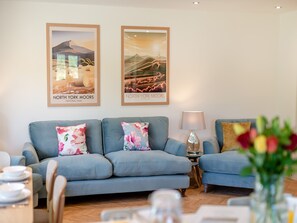 A living room with a blue sofa set, two framed posters of North York Moors, a lamp, and a vase with flowers.