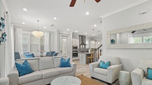 Una sala de estar moderna con un sofá gris, dos sillones y una mesa de centro redonda. Se ve una cocina al fondo con armarios blancos y una zona de comedor.