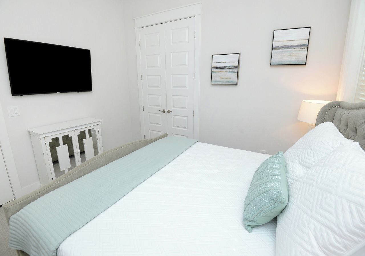 Ein Schlafzimmer mit einem Bett, einem an der Wand montierten Fernseher, einer Tür und einem gerahmten Bild.