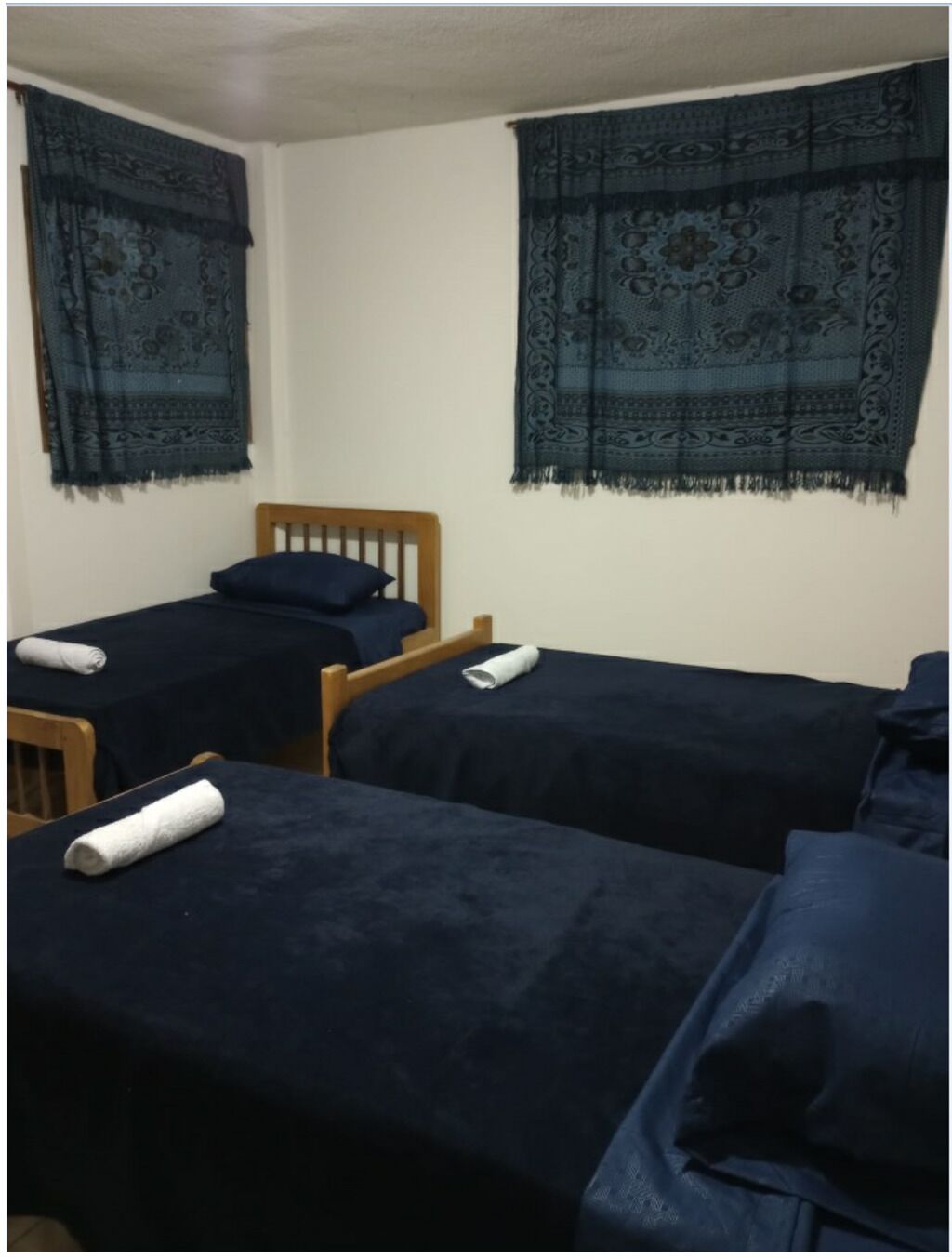 A dormitory room with two single beds, each with a dark blue bedspread and a rolled white towel. The room has a wooden headboard and two windows with dark blue curtains featuring intricate patterns.