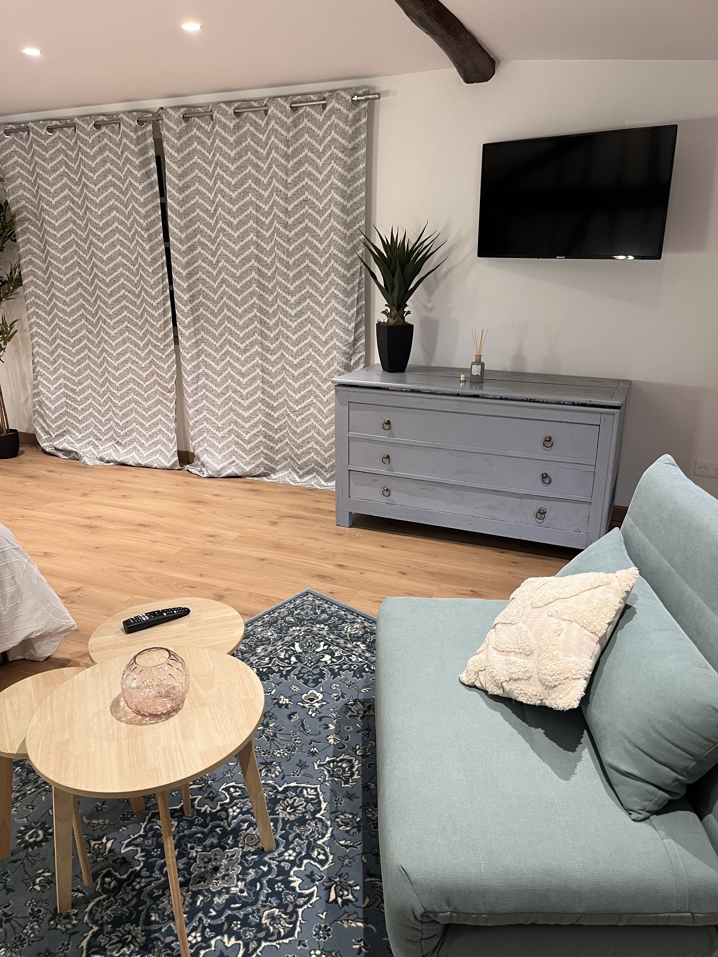 Een woonkamer met een grijze bank, een klein rond tafeltje, een aan de muur bevestigde televisie en een grijze kast.