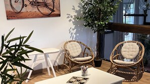 Un salon chaleureux avec une table basse blanche, deux fauteuils en osier, une grande plante en pot et un tableau représentant un vélo accroché au mur.