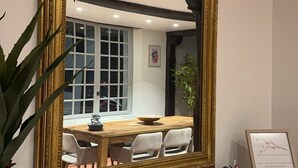 Une pièce comprenant un grand miroir décoratif, une table en bois, des chaises, une plante en pot, une horloge et un certificat encadré posé sur la commode.