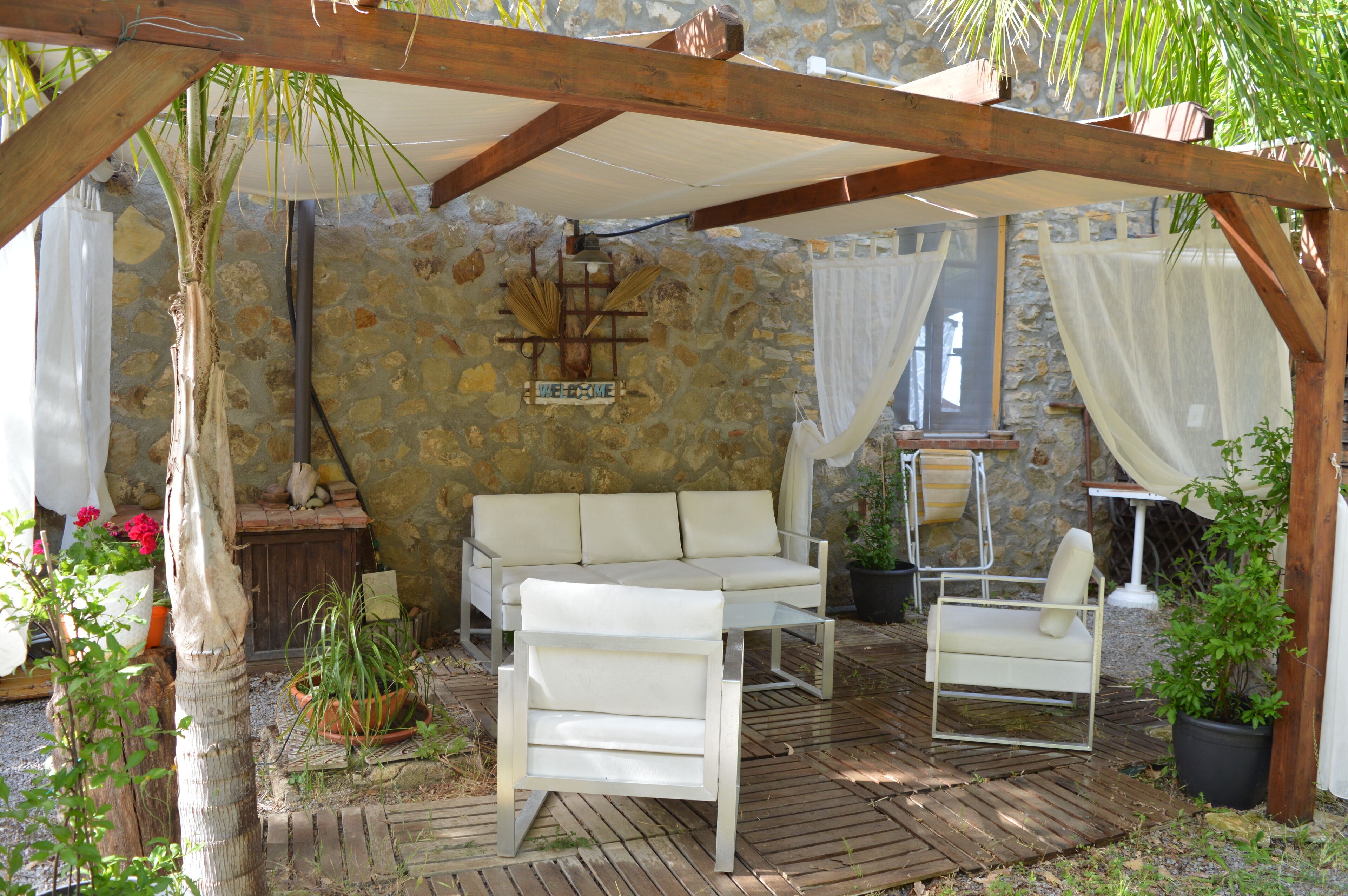 Un espacio de estar al aire libre techado, con pared de piedra, pérgola de madera y plantas en macetas.