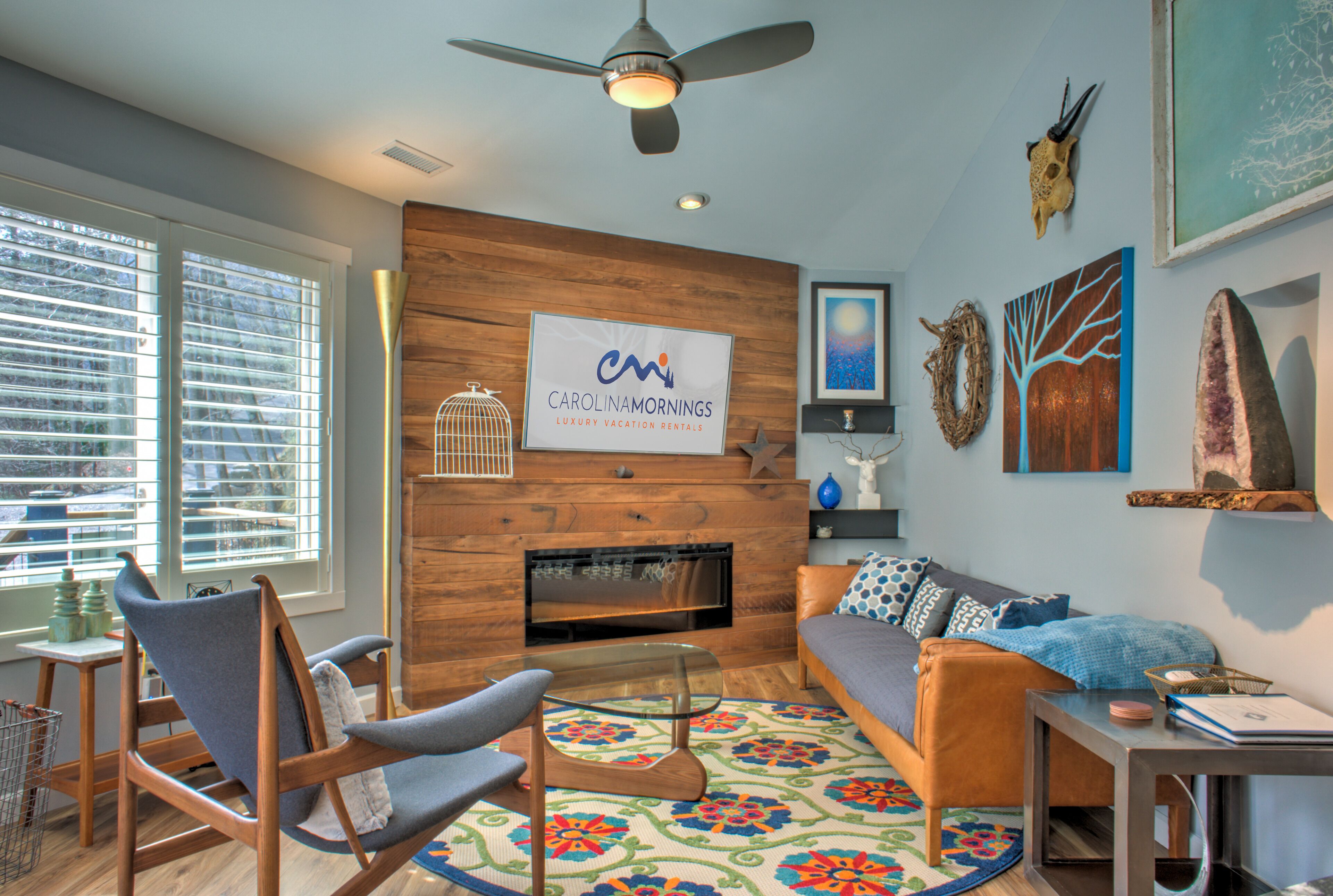 A modern living room with a fireplace, a ceiling fan, and a sign for Carolinamornings Luxury Vacation Rentals.